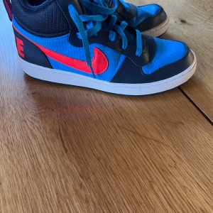 Blå och svarta Nike sneakers - Säljer ett par coola Nike sneakers i blått och svart med röda detaljer. Skorna har en klassisk design med snörning och en bekväm sula. Perfekta för vardagsbruk eller träning. De är i bra skick och redo för nya äventyr!