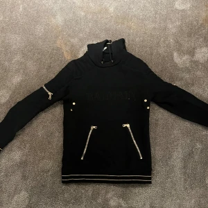 Svart hoodie från Balmain - Säljer en snygg svart hoodie från Balmain med coola dragkedjedetaljer på fickorna och ärmarna. Den har en quiltad design på axlarna och en hög krage med snörning. Perfekt för höst och vinter, både stilren och bekväm. Passar lika bra till vardags som till fest!
