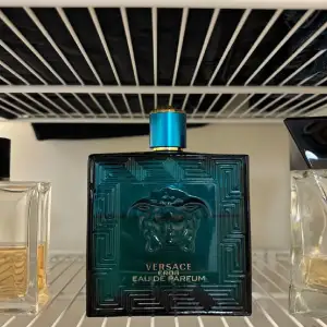 Säljer en flaska Versace Eros Eau de Parfum. Flaskan är fyrkantig och har en djup turkos färg med ett elegant mönster och Versaces ikoniska Medusa-logga på framsidan. Korken är också turkos med en gyllene kant. Perfekt för den som vill ha en doft som utstrålar självsäkerhet och stil.