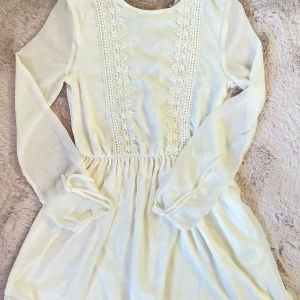 Vit klänning med spetsdetaljer - Superfin vit klänning med spetsdetaljer och snörning på baksidan. Den har långa ärmar och en söt, feminin stil. Perfekt för vår och sommar, kanske till en fest eller midsommarfirande. Materialet känns lätt och luftigt, vilket gör den bekväm att bära hela dagen.