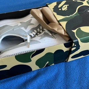 Vita och grå sneakers från A Bathing Ape - Säljer ett par snygga vita och grå sneakers från A Bathing Ape. Skorna är i nytt skick. De kommer i en cool kamouflagemönstrad låda. Perfekta för att ge din outfit en streetwear-känsla!