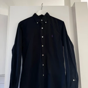 Svart skjorta från Ralph Lauren - Säljer en stilren svart skjorta från Ralph Lauren i slim fit. Skjortan har långa ärmar och klassiska knappar framtill. Perfekt för både vardag och festliga tillfällen. Den är i mycket bra skick och redo för en ny ägare!