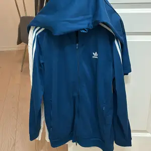 Fin blå Adidas tracksuit, köpt på Adidas i Spanien. Tracksuit; dvs både koftan/tröjan och byxorna ingår. Fint skick på dem, alltid tvättade enligt anvisningar. Färg: Blå Storlek: L på tröjan & XL på byxorna Byxorna passar dock även L då de spänns åt