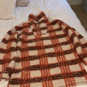 Rutig teddyjacka från Missguided - Supermysig teddyjacka från Missguided i rutig design med bruna och orangea toner. Perfekt för höst och vinter med sin varma och gosiga känsla. Jackan har en dragkedja framtill och långa ärmar. Perfekt för att hålla stilen när det är kallt ute!