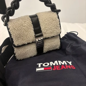 Beige och svart axelväska från Tommy Jeans - Säljer en superfin beige och svart axelväska från Tommy Jeans. Väskan har ett fluffigt teddy-material och en cool kedjedetalj. Perfekt för höst och vinter! Den är i nyskick och kommer med en justerbar axelrem.