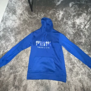 Blå hoodie och byxor från Trapstar - Säljer en snygg blå hoodie och matchande byxor från Trapstar. Hoodien har en cool text på framsidan och en klassisk känguruficka. Byxorna är bekväma med elastisk midja och samma textdetalj. Perfekt för en avslappnad stil eller streetwear-look. Passar både höst och vår!
