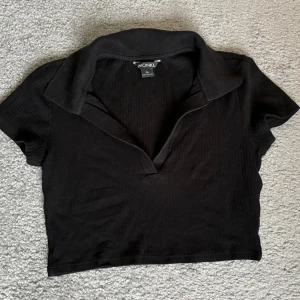 Svart ribbad topp från Monki - Säljer en svart ribbad topp från Monki i storlek XS. Den har en klassisk krage och korta ärmar, perfekt för både vardag och fest. Toppen är i bra skick och passar till det mesta i garderoben. 🖤
