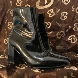 Svarta lackade stövletter med klack - Snygga svarta stövletter i lackat material från Gina tonic. De har en elegant spetsig tå och en stabil klack som ger en chic look. Perfekta för både fest och vardag. Dragkedja på sidan för enkel på- och avtagning. Storlek 37.