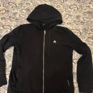 Svart hoodie från Moose Knuckles - Säljer en svart hoodie från Moose Knuckles i bra skick. Den har en dragkedja framtill och en liten logga på bröstet.. Ny pris 3000kr på johnells kungens kurva