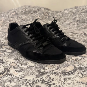 Svarta sneakers från Boss - Säljer ett par stilrena svarta sneakers från Boss. De har en snygg kombination av skinn och mocka med klassisk snörning. Perfekta för både vardag och fest! Skorna är i bra skick utan att det finns lite slitage på insidan av skon se 3 bilden