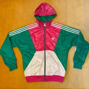 Cool Adidas vindjacka stl M - Klassisk Adidas jacka i härliga färger, storlek M. I ok skick, del av söm gått upp. Huva och två fickor med dragkedjor.