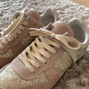 Glittriga sneakers i rosa mocka - Säljer ett par snygga sneakers i rosa mocka med glittriga detaljer. Skorna har snörning och en bekväm passform, perfekta för både vardag och fest. De är i bra skick och redo för nya äventyr!