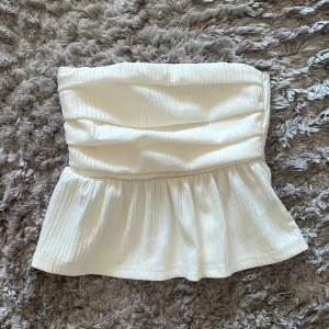 Vit axelbandslös topp - Säljer en superfin vit axelbandslös topp med en smickrande passform. Toppen har en volangdetalj nedtill som ger en söt touch. Perfekt för sommarens alla fester eller en dag på stan. Materialet känns mjukt och bekvämt mot huden.