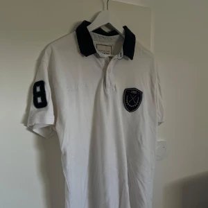 Vit pikétröja med broderad logga - Säljer en snygg vit pikétröja från Polo Club. Den har en broderad logga på bröstet och siffran 8 på ärmen. Kragen är mörkblå vilket ger en fin kontrast. Perfekt för en avslappnad men stilren look!