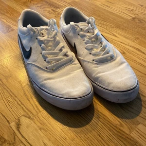 Vita Nike sneakers med svart Swoosh - Säljer ett par vita Nike sneakers med den klassiska svarta Swoosh-loggan på sidan. Skorna har snörning och en bekväm passform. De är i välanvänt skick men fortfarande snygga och funktionella. Perfekta för vardagsbruk!