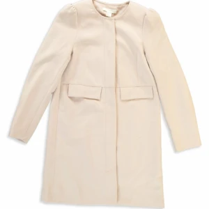Beige kappa - Säljer en stilren beige kappa med långärmad design. Perfekt för höst och vår, med en klassisk look som passar till alla tillfällen. Kappan har en enkel och elegant stil utan krage och med diskreta detaljer framtill. Den har en gömd dragkedja. 