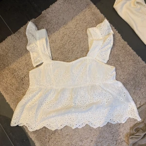 Vit blus med spetsdetaljer - Säljer en superfin vit blus med spetsdetaljer och volanger på axlarna. Perfekt för sommardagar eller en festlig kväll. Den har en luftig passform och är tillverkad i ett lätt och bekvämt material. Passar perfekt till jeans eller kjol!