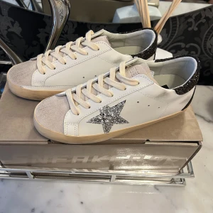 Glittriga sneakers från Golden Goose - Säljer ett par snygga vita sneakers från Golden Goose med glittriga detaljer. Skorna har en glittrig stjärna på sidan och glittriga hälpartier. De är i mycket bra skick och perfekta för att ge din outfit lite extra bling. Skorna har snörning och en bekväm passform. Kommer med dustbag och vykort från Golden goose./Dma