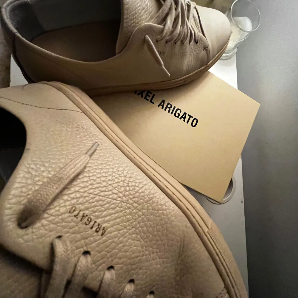 Säljer ett par snygga beige sneakers från Axel Arigato i mycket bra skick. De är gjorda i skinn med en stilren design och har snörning framtill. Perfekta för både vardag och fest! 🕺 Nypris 2700kr pris kan möjligtvis sänkas.. Skor.