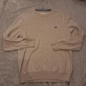 Beige tröja från GANT - Säljer en stilren beige tröja från GANT i mycket bra skick. Den har en broderad logga på bröstet och är tillverkad i ett mjukt material som känns skönt mot huden. Perfekt för både vardag och lite finare tillfällen. Passar bra till höst och vår! Säljer den för 399kr