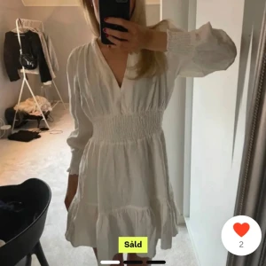 Vit klänning med volangdetaljer - Säljer en superfin vit klänning med v-ringning och långa ärmar. Den har en smockad midja som ger en snygg passform och volangdetaljer längst ner. Perfekt för sommarens alla fester eller en mysig vårdag. Klänningen är i mycket bra skick och passar perfekt för en stilren look.