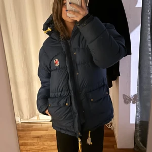 Blå dunjacka från Fjällräven - Säljer en blå dunjacka från Fjällräven i mycket bra skick förutom en defekt vid fickan som går lätt o laga!! Jackan har en mysig huva och praktiska fickor med knappar. Perfekt för kalla vinterdagar. Den har en klassisk Fjällräven-logga på bröstet och stängs med dragkedja och knappar. Supervarm och skön! (Lägsta pris som accepteras är 3200) 