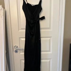 Svart siden långklänning från Tommy Hilfiger - Elegant svart långklänning från Tommy Hilfiger i storlek EUR 32. Klänningen har en omlott-design med knytband i midjan och smala axelband och är i siden material. 