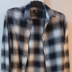 Rutig skjorta från H&M - Snygg rutig skjorta från H&M i grått och vitt. Perfekt för både vardag och fest! Skjortan har långa ärmar och knappar framtill. Den är gjord i ett mjukt och bekvämt material som känns skönt mot huden. Passar bra till jeans eller chinos för en avslappnad look.
