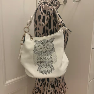 Vit handväska med ett uggletryck  - Säljer den här supergulliga vita handväskan med prydnadstenar formade till uggla. Mycket bra kvalitet då den nästan aldrig blivit använd. Finns ett oöppnat axelremsband så man kan välja om man vill ha en handväska eller axelremsväska! Bredden är 25 cm och höjden är 24-27 cm för att väskan böjd.💕