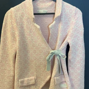 Rosa stickad jacka från Odd Molly - Superfin rosa stickad jacka från Odd Molly med ett diskret mönster och broderad detalj på ärmen. Den har en söt knytning framtill och två fickor med knappar. Perfekt för vår och höst när du vill ha något mysigt och stiligt på samma gång!