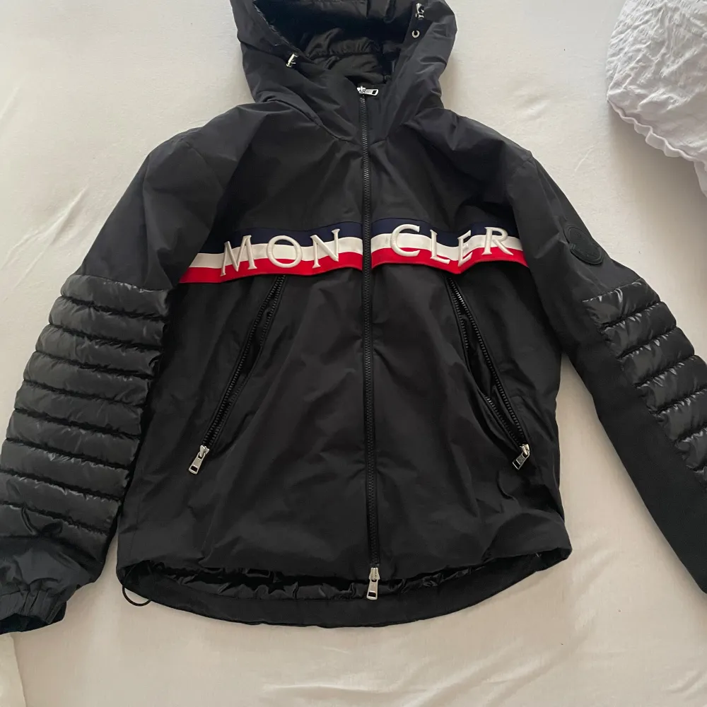 Säljer en snygg svart Moncler jacka i mycket bra skick exakt som ny. Den är slutsåld överallt och artister som sarettii och dizzy har denna modell. Den har en cool design med Moncler-logga över bröstet och röd-vit rand. Jackan är quiltad och har dragkedja framtill samt på fickorna. Perfekt för höst och vår med sin stilrena look och bekväma passform. Passar både till vardags och fest!. Jackor.