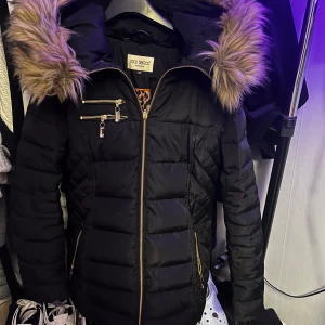 Svart pufferjacka med pälsdetaljer från Roco Baroco - Säljer en snygg svart pufferjacka från Roco Baroco i storlek 36. Jackan har en mysig pälsdetalj på huvan och ett coolt leopardmönstrat foder. Den har dragkedja och flera praktiska fickor med dragkedjor. Perfekt för kalla vinterdagar! Har dock håll i fickan men går bra att fixa