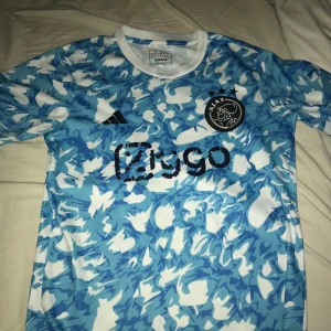 Blå och vit Ajax fotbollströja från Adidas - Säljer en snygg Ajax fotbollströja i blått och vitt från Adidas. Tröjan har korta ärmar och ett coolt mönster med klubbens logga och sponsortryck på framsidan. Perfekt för alla Ajax-fans! 🏆passar även M
