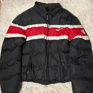 Svart och röd pufferjacka från No Fear - Säljer en cool svart pufferjacka från No Fear med röda och vita ränder. Jackan har dragkedja och broderade detaljer på bröstet och ärmen. Perfekt för vintern med sin varma och puffiga stil. Passar både till vardags och sportiga aktiviteter.