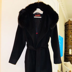 Svart kappa från Max Mara - Säljer en elegant svart kappa från Max Mara i mycket bra skick. Den har en lyxig pälsdetalj runt kragen och ett knytbälte i midjan för en snygg passform. Perfekt för höst och vinter, med långa ärmar och en klassisk stil. Passar både till vardags och festliga tillfällen. Storlek 34.