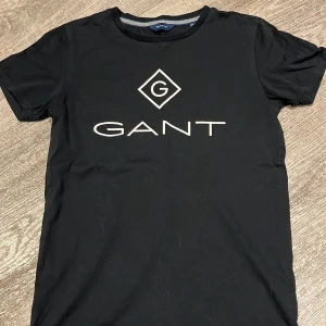 Svart t-shirt från GANT - Snygg svart t-shirt från GANT i bomull med deras ikoniska logga tryckt på framsidan. Perfekt för en casual look, oavsett om du ska på stan eller hänga med kompisar. T-shirten har en normal passform och korta ärmar. Ett måste i garderoben för alla som gillar stilren design!