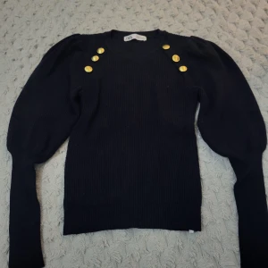 Svart ribbad tröja från Zara - Säljer en snygg svart ribbad tröja från Zara med guldknappar på axlarna. Tröjan har långa ärmar och en klassisk rund halsringning. Perfekt för både vardag och fest! 🖤