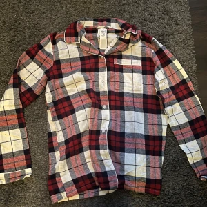 Rutig flanellskjorta - Säljer en mysig rutig flanellskjorta i rött, svart och vitt. Skjortan har långa ärmar och knappar framtill. Perfekt för höst och vinter, och den är i nyskick. Prislappen finns kvar och den är aldrig använd. Pyjamasskjorta i storlek 170