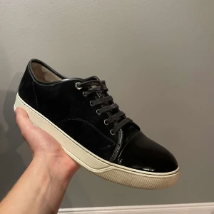 Svarta sneakers från Lanvin - Säljer ett par stilrena svarta sneakers från Lanvin i mycket bra skick. Skorna har en snygg kombination av mocka och skinn med en vit sula. Perfekta för både vardag och fest! Kommer med originalpåse.