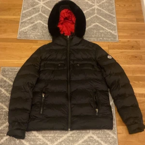 Svart dunjacka från Moncler - Säljer en snygg svart dunjacka från Moncler i mycket bra skick. Jackan har en röd insida och en mysig huva med päls. Den är perfekt för vintern och har praktiska dragkedjor på fickorna. Passar både till vardags och festliga tillfällen. Storlek L men passar perfekt till ändå jag har använt den och har M