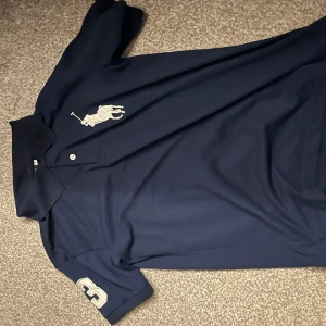 Mörkblå pikétröja med broderad logga - Säljer en snygg mörkblå pikétröja från Ralph Lauren i storlek M. Tröjan har en stor broderad logga på bröstet och siffran 3 på ärmen. Perfekt för en stilren och sportig look. Passar både till vardags och fest!