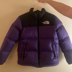 Lila dunjacka från The North Face - Säljer en supervarm och snygg lila dunjacka från The North Face. Jackan har svarta detaljer och en dragkedja framtill. Perfekt för kalla vinterdagar med sin 700-fill power. Den är i mycket bra skick med en defekt, se bild!