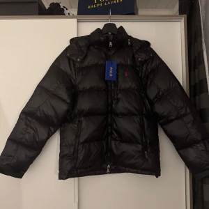 Svart dunjacka från Polo Ralph Lauren - Säljer en snygg svart dunjacka från Polo Ralph Lauren i mycket bra skick. Jackan har en broderad röd logga på bröstet och praktiska dragkedjor. Perfekt för kalla vinterdagar med sin pufferstil och långa ärmar. Huvan ger extra skydd mot väder och vind. Passar både till vardags och lite finare tillfällen.