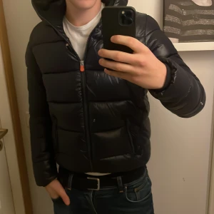 Svart pufferjacka från Save the Duck - Säljer en svart pufferjacka från Save the Duck i mycket bra skick. Jackan är 100% animal free och har en dragkedja framtill. Perfekt för höst och vinter med sin varma och stilrena design. Den har en normal passform och är långärmad. Huvan ger extra skydd mot kyla. Passar både till vardags och lite finare tillfällen. 187cm lång på bilden!