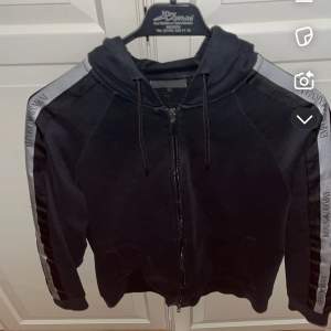 Säljer en svart hoodie från Armani Exchange i bra skick. Den har en dragkedja framtill och långa ärmar med gråa detaljer längs sidorna. Perfekt för höst och vår, med en bekväm passform och huva med dragsko.