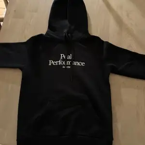 Säljer en svart hoodie från Peak Performance med vit text på bröstet. Den har en klassisk design med en stor ficka fram och justerbar huva. Perfekt för kyliga dagar eller som en stilren vardagströja. Passar både till skolan och fritiden!      OBS! Barnstorlek 