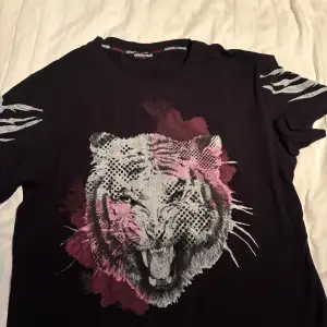 Säljer en cool svart t-shirt från Roberto Cavalli med ett stort tigertryck i vitt och rosa på framsidan. T-shirten har korta ärmar och är perfekt för en avslappnad stil. Den är gjord av bomull och elastan för en bekväm passform. Roberto Cavalli-loggan finns på baksidan.