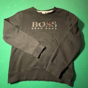 Svart tröja från Hugo Boss - Säljer en stilren svart tröja från Hugo Boss i storlek 160. Tröjan har långa ärmar och ett klassiskt tryck med BOSS-loggan på bröstet. Perfekt för både vardag och festliga tillfällen. Skön och enkel att matcha med det mesta i garderoben! Priset är verkligen inte hugget i sten!