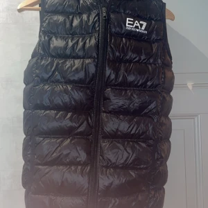 Svart dunväst från Emporio Armani - Säljer en snygg svart dunväst från Emporio Armani. Västen har en dragkedja framtill och är quiltad för extra värme. Perfekt för höst och vår när du vill hålla dig varm men ändå se stilren ut. Passar både till vardags och festliga tillfällen. Storlek: passar dig som är 173-178 tveka inte att höra av dig!