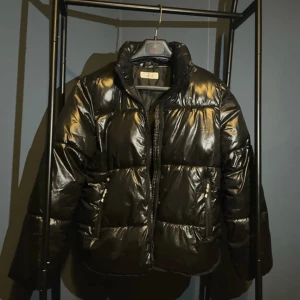 Svart pufferjacka från H&M - Säljer en snygg svart pufferjacka från H&M i storlek M. Jackan är perfekt för kalla dagar med sitt blanka material och quiltade design. Den har en dragkedja framtill och långa ärmar. Perfekt för vintern och hösten!